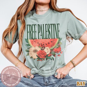 May include: Sage green t-shirt with the text "FREE PALESTINE" in dark green. The shirt features a watermelon slice and floral design in red, white, and beige. The shirt is knotted at the waist.