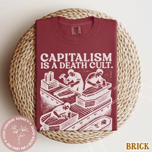 K&ouml;nnte beinhalten: Ziegelrotes T-Shirt mit dem Text "CAPITALISM IS A DEATH CULT." und einer Grafik von Personen, die S&auml;rge bauen. Das Shirt ist auf einer gewebten, kreisf&ouml;rmigen Oberfl&auml;che gefaltet. Comfort Colors Marke.