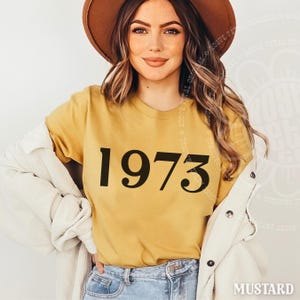 May include: A mustard yellow short-sleeved t-shirt featuring the black text 1973 printed in a serif font on the front. The shirt is worn under a white corduroy button-down jacket, paired with blue denim jeans and a brown wide-brimmed hat.