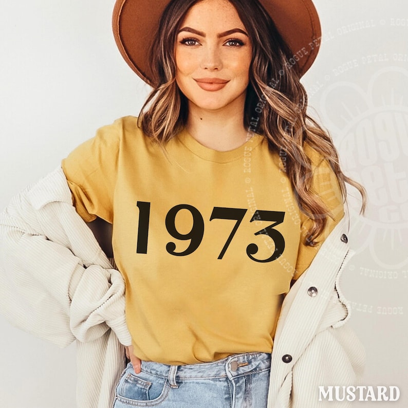 May include: A mustard yellow short-sleeved t-shirt featuring the black text 1973 printed in a serif font on the front. The shirt is worn under a white corduroy button-down jacket, paired with blue denim jeans and a brown wide-brimmed hat.