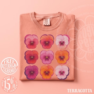 Puede incluir: Una camiseta de color terracota con una cuadr&iacute;cula de nueve ilustraciones de flores de pensamiento en tonos rosa, rojo y naranja. La prenda tiene cuello redondo y una etiqueta de la marca Comfort Colors en el interior. La palabra Terracotta aparece abajo.