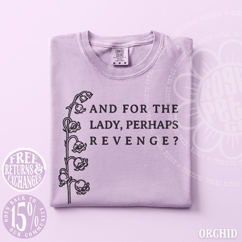以下が含まれることがあります： 左側にスズランの茎の黒いグラフィック、右側に「AND FOR THE LADY, PERHAPS REVENGE?」という黒い文字がプリントされたライトパープルのTシャツ。クルーネックで、首元にはComfort Colorsのブランドラベルが付いています。