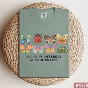 May include: Sage green t-shirt with a graphic of colorful butterflies and a ladybug. The text "All of us different. None of us less." is printed on the shirt.