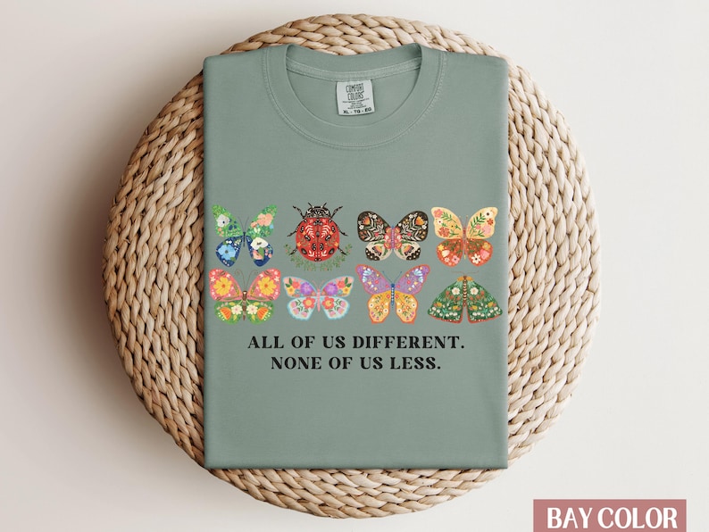 May include: Sage green t-shirt with a graphic of colorful butterflies and a ladybug. The text "All of us different. None of us less." is printed on the shirt.