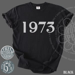 May include: A black short-sleeved t-shirt featuring the number 1973 printed in a large, light gray serif font across the chest. The garment has a crew neckline and a standard fit, with a small white brand label visible inside the collar area.