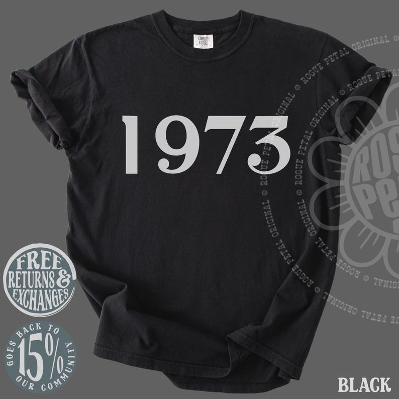 May include: A black short-sleeved t-shirt featuring the number 1973 printed in a large, light gray serif font across the chest. The garment has a crew neckline and a standard fit, with a small white brand label visible inside the collar area.