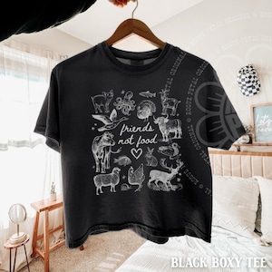 Puede incluir: Camiseta negra con un gr&aacute;fico blanco de varios animales y el texto "friends not food". La camiseta est&aacute; colgada de una percha de madera.