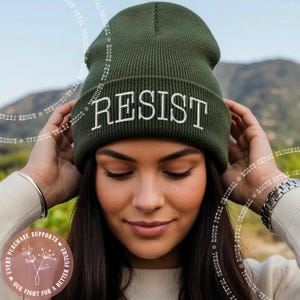 May include: An olive green knit beanie with the word "RESIST" embroidered in white across the front. The beanie is worn by a person. The image also includes the text "EVERY PURCHASE SUPPORTS OUR FIGHT FOR A BETTER FUTURE".
