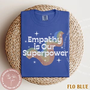 Puede incluir: Una camiseta azul con el texto blanco "Empathy is Our Superpower" y un dise&ntilde;o de arco&iacute;ris. La camiseta est&aacute; doblada sobre una superficie circular tejida. Tambi&eacute;n es visible la frase "Every purchase supports our fight for a better future".