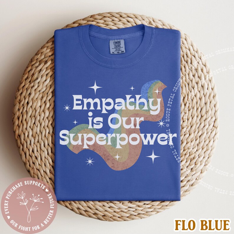Puede incluir: Una camiseta azul con el texto blanco "Empathy is Our Superpower" y un dise&ntilde;o de arco&iacute;ris. La camiseta est&aacute; doblada sobre una superficie circular tejida. Tambi&eacute;n es visible la frase "Every purchase supports our fight for a better future".