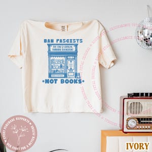 May include: Ivory-colored t-shirt with the text "BAN FASCISTS" above a blue illustration of a bookstore. The text "NOT BOOKS" is below the bookstore graphic. The shirt also has the text "THE EVIL CRITICAL THINKING BOOKSTORE".