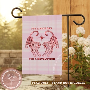 May include: A pink garden flag with a plaid pattern features two red leopard illustrations facing each other. The text reads "IT'S A NICE DAY FOR A REVOLUTION". The flag is displayed on a black metal stand.