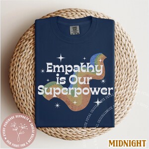 Puede incluir: Una camiseta azul marino con el texto "Empathy is Our Superpower" en blanco, con un gr&aacute;fico de arco&iacute;ris. La camiseta est&aacute; doblada sobre una estera circular tejida. La palabra "Midnight" est&aacute; en la parte inferior.