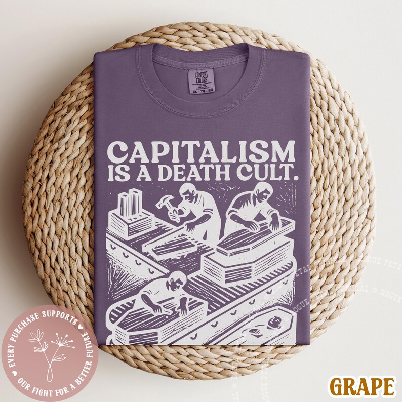 K&ouml;nnte beinhalten: Ein lilafarbenes T-Shirt mit dem wei&szlig;en Text "CAPITALISM IS A DEATH CULT." und einer Grafik von Arbeitern, die S&auml;rge bauen. Das T-Shirt ist auf einer gewebten Matte gefaltet. Ein kleiner runder Aufkleber mit Text ist sichtbar.