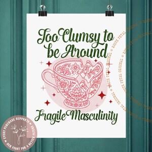 May include: A white poster with the text "Too Clumsy to be Around" and "Fragile Masculinity" in green and black. A pink teacup design is in the center, with cracks and floral patterns. The poster is held by two black clips.