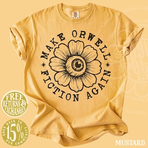 May include: A mustard yellow short-sleeve t-shirt featuring a black graphic print of a flower with an eye in the center. The text "MAKE ORWELL FICTION AGAIN" is printed in a circular pattern around the flower. The shirt has a crew neckline.
