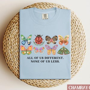 May include: Light blue t-shirt with a graphic of colorful butterflies and a ladybug. The text "All of us different. None of us less." is printed on the shirt.