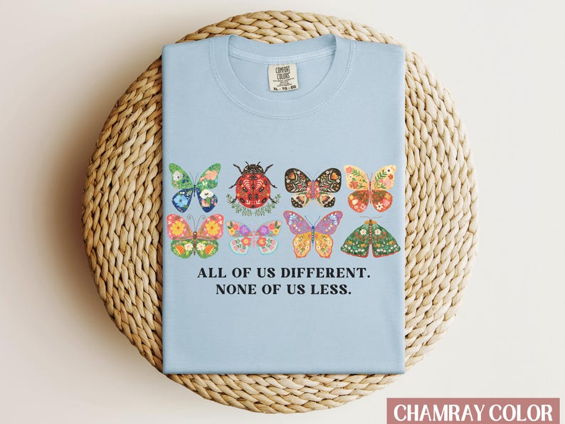 May include: Light blue t-shirt with a graphic of colorful butterflies and a ladybug. The text "All of us different. None of us less." is printed on the shirt.
