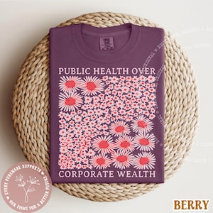 May include: Berry-colored t-shirt with the text "PUBLIC HEALTH OVER CORPORATE WEALTH" and a floral design. The design features white and red daisies. The shirt is displayed on a woven, circular mat.
