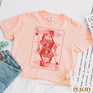 以下が含まれることがあります： 桃色のTシャツに、ハートの女王のトランプのグラフィックがプリントされています。女王はサングラスをかけて、花を持っています。