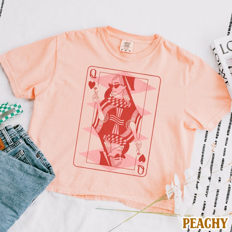 以下が含まれることがあります： 桃色のTシャツに、ハートの女王のトランプのグラフィックがプリントされています。女王はサングラスをかけて、花を持っています。
