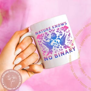 May include: White ceramic mug with the text "NATURE KNOWS NO BINARY" in pink and blue. The mug features a floral and bird design in the same colors. The mug is held against a pink background.