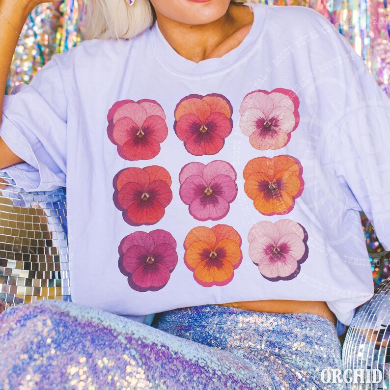 Puede incluir: Una camiseta blanca de manga corta con una cuadr&iacute;cula de nueve flores de pensamiento en varios tonos de rosa, rojo, naranja y morado. El gr&aacute;fico floral est&aacute; dispuesto en tres filas de tres, mostrando p&eacute;talos distintos y centros oscuros.