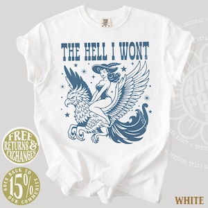 May include: A white t-shirt featuring a blue graphic print of a person wearing a wide-brimmed hat riding a large winged creature. The text "THE HELL I WONT" is printed in blue above the illustration, which is accented with small star shapes.