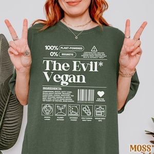May include: A dark green t-shirt with white text that reads "100% Plant-Powered 0% Regrets The Evil* Vegan". The shirt also features a barcode, a heart with the text "For the Voiceless", and a series of icons representing animal lover, planet activist, gourmet foodie, half human half tofu, and vegan spot gps.