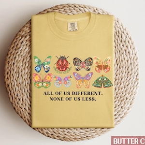 May include: A yellow t-shirt with a graphic of colorful butterflies and a ladybug. The text "All of us different. None of us less." is printed on the shirt.