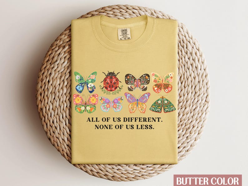 May include: A yellow t-shirt with a graphic of colorful butterflies and a ladybug. The text "All of us different. None of us less." is printed on the shirt.