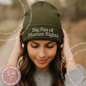 May include: Olive green beanie with the embroidered text "Big Fan of Human Rights" in white. The hat is ribbed and has a folded brim. The image also includes a small round sticker with text and a floral design.