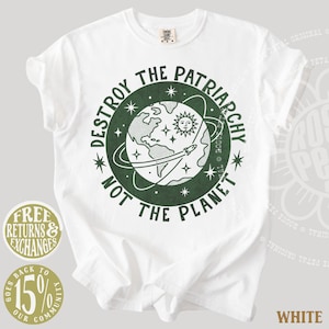 May include: A white short-sleeve t-shirt featuring a dark green circular graphic on the chest. The design depicts a globe, a sun, stars, and a rocket ship, surrounded by the text "DESTROY THE PATRIARCHY NOT THE PLANET" in a bold, dark green font.