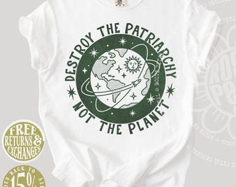 Puede incluir: Una camiseta blanca de manga corta con un gráfico circular de color verde oscuro en el pecho. El diseño muestra un globo terráqueo, un sol, estrellas y un cohete, rodeados por el texto "DESTROY THE PATRIARCHY NOT THE PLANET" en letra verde oscuro.