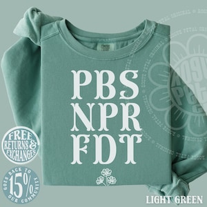 May include: A light green crewneck sweatshirt featuring white text that reads PBS, NPR, FDT in a stacked serif font. Below the text is a small white line-art illustration of three flowers. The garment has a ribbed collar and long sleeves.