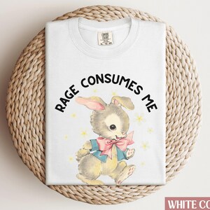 Op de afbeelding: Wit T-shirt met een cartoonkonijn met een blauwe strik en de tekst "RAGE CONSUMES ME".
