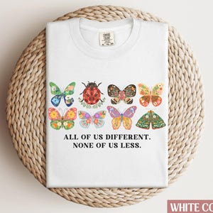 May include: White t-shirt with a graphic of colorful butterflies and a ladybug. The text "All of us different. None of us less." is printed below the graphic.