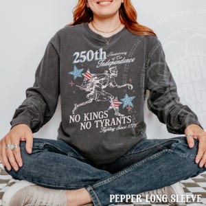 May include: Dark gray long-sleeve shirt with a graphic commemorating the 250th anniversary of the Independence. The design includes stars, a running figure, and the text "No Kings No Tyrants Fighting Since 1776".