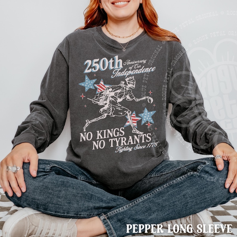 May include: Dark gray long-sleeve shirt with a graphic commemorating the 250th anniversary of the Independence. The design includes stars, a running figure, and the text "No Kings No Tyrants Fighting Since 1776".