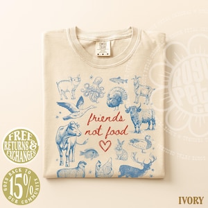Puede incluir: Camiseta de color marfil con una ilustraci&oacute;n azul de varios animales y el texto rojo "friends not food". El dise&ntilde;o incluye un s&iacute;mbolo de coraz&oacute;n. La camiseta tiene una etiqueta Comfort Colors.