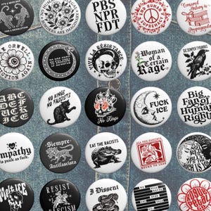 May include: Collection of round buttons with various designs and text. Designs include a skull, a cat, a snake, a crocodile, and phrases like "Eat the Racists" and "No Kings." The buttons are in black, white, and red.