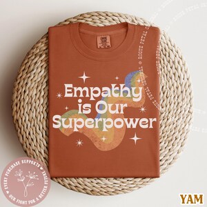 Puede incluir: Una camiseta de color &oacute;xido con el texto "Empathy is Our Superpower" en blanco, con un dise&ntilde;o de arco&iacute;ris retro. La camiseta est&aacute; doblada sobre una cesta tejida. La camiseta tiene una etiqueta que dice "Comfort Colors".