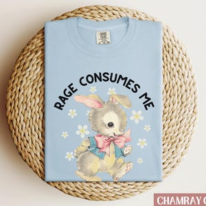 Op de afbeelding: Een lichtblauw t-shirt met een cartoonkonijn dat een blauwe jas en een roze strik draagt. De tekst "RAGE CONSUMES ME" is in zwart op het shirt gedrukt.