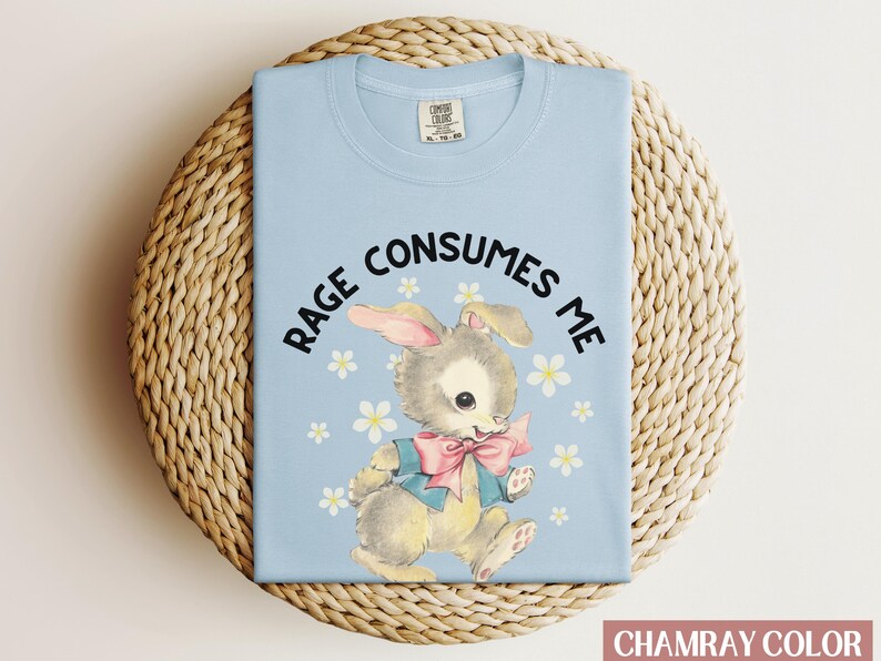 Op de afbeelding: Een lichtblauw t-shirt met een cartoonkonijn dat een blauwe jas en een roze strik draagt. De tekst "RAGE CONSUMES ME" is in zwart op het shirt gedrukt.
