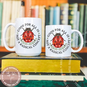 May include: Two white ceramic mugs with the text "Good for All Is Not a Radical Concept" and a red ladybug design. The mugs are on a stack of books with a blurred bookshelf background. The ladybug has a detailed pattern.