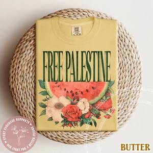 May include: A yellow t-shirt with the text "FREE PALESTINE" in green, above a watermelon slice and floral arrangement. The shirt is folded on a woven surface. The word "BUTTER" is at the bottom right.
