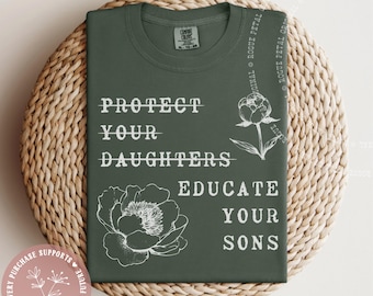 May include: A moss green t-shirt with the text "PROTECT YOUR DAUGHTERS EDUCATE YOUR SONS" in white. The shirt features floral illustrations and a round sticker with text. The shirt is folded on a woven mat.