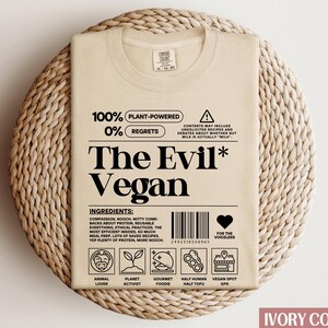 May include: A beige t-shirt featuring a graphic design that reads "100% Plant-Powered 0% Regrets The Evil* Vegan". The graphic includes a barcode, a heart with the text "For the voiceless", and icons representing animal lover, planet activist, gourmet foodie, half human half tofu, and vegan spot GPS.
