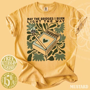 May include: Mustard-colored t-shirt with the text "MAY THE BRIDGES I BURN LIGHT THE WAY." The graphic features a matchbox with a heart and a lit match, surrounded by green floral elements. The shirt has rolled sleeves.