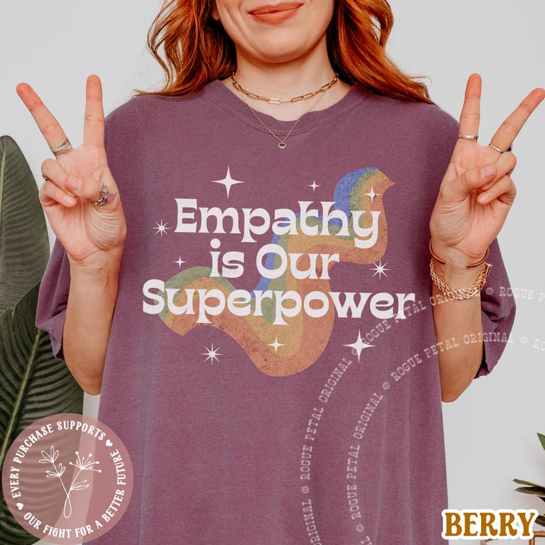 Puede incluir: Camiseta de color bayas con el texto "Empathy is Our Superpower" en blanco y un dise&ntilde;o de arco&iacute;ris. La camiseta tiene un corte holgado y est&aacute; hecha de un material suave. La camiseta tambi&eacute;n tiene el texto "ROGUE PETAL ORIGINAL" impreso en el lateral.